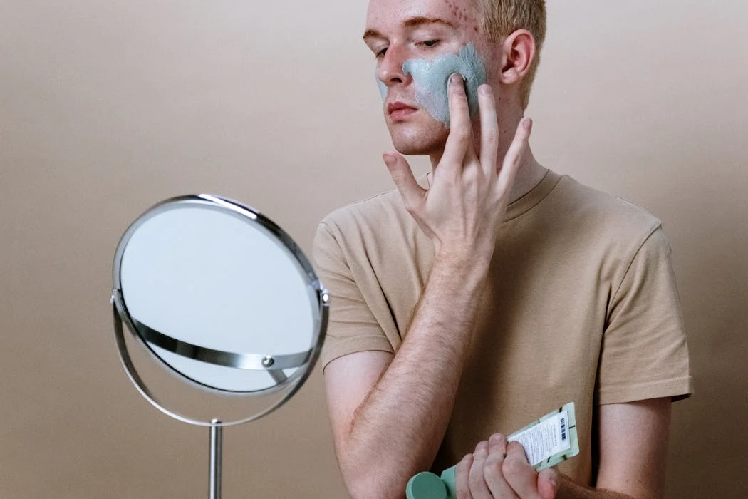 Young man applying clay mask for skincare routine indoors with a mirror.